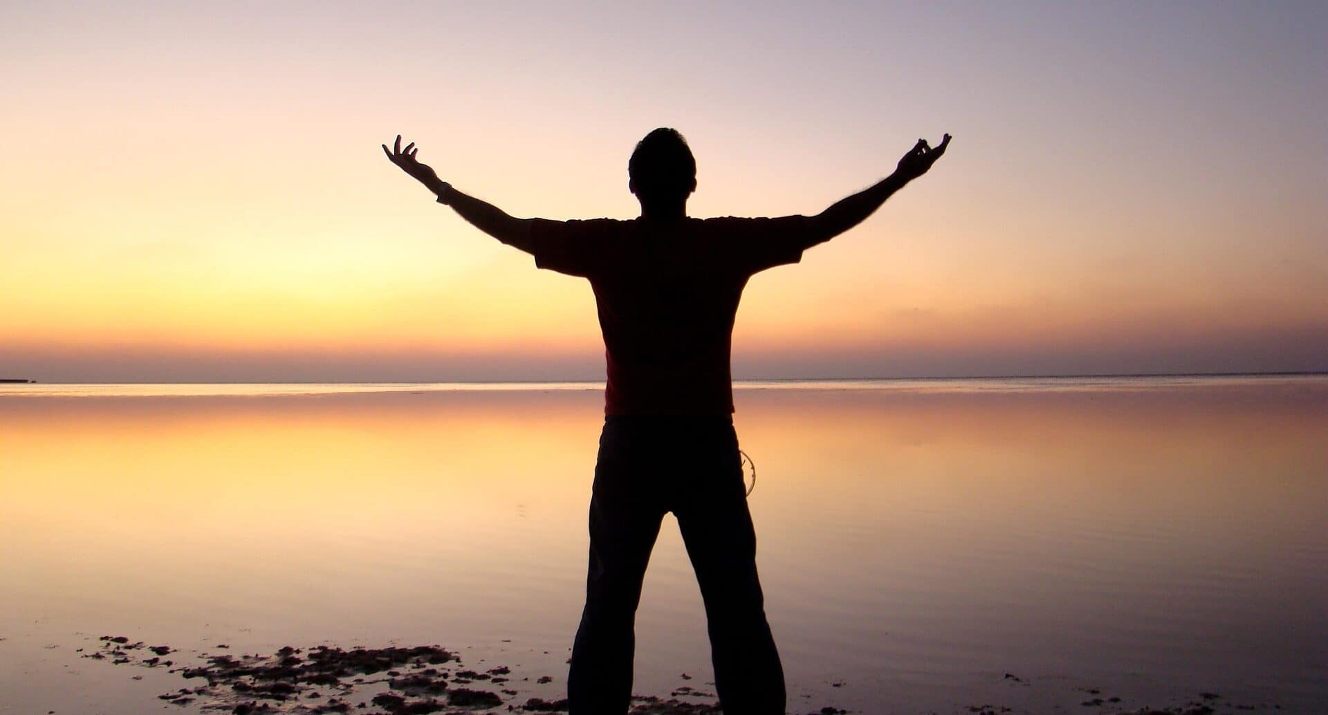 A silhouette of a man with his hands raised, against the sea and a clear sunset sky in the background